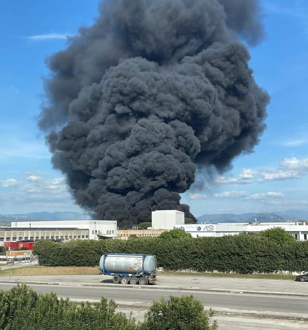 [Nessun titolo dell'immagine] - Un denso fumo nero si alza nel cielo a causa di un incendio in uno stabilimento industriale, con un'autocisterna su una strada vicina e degli edifici in primo piano.